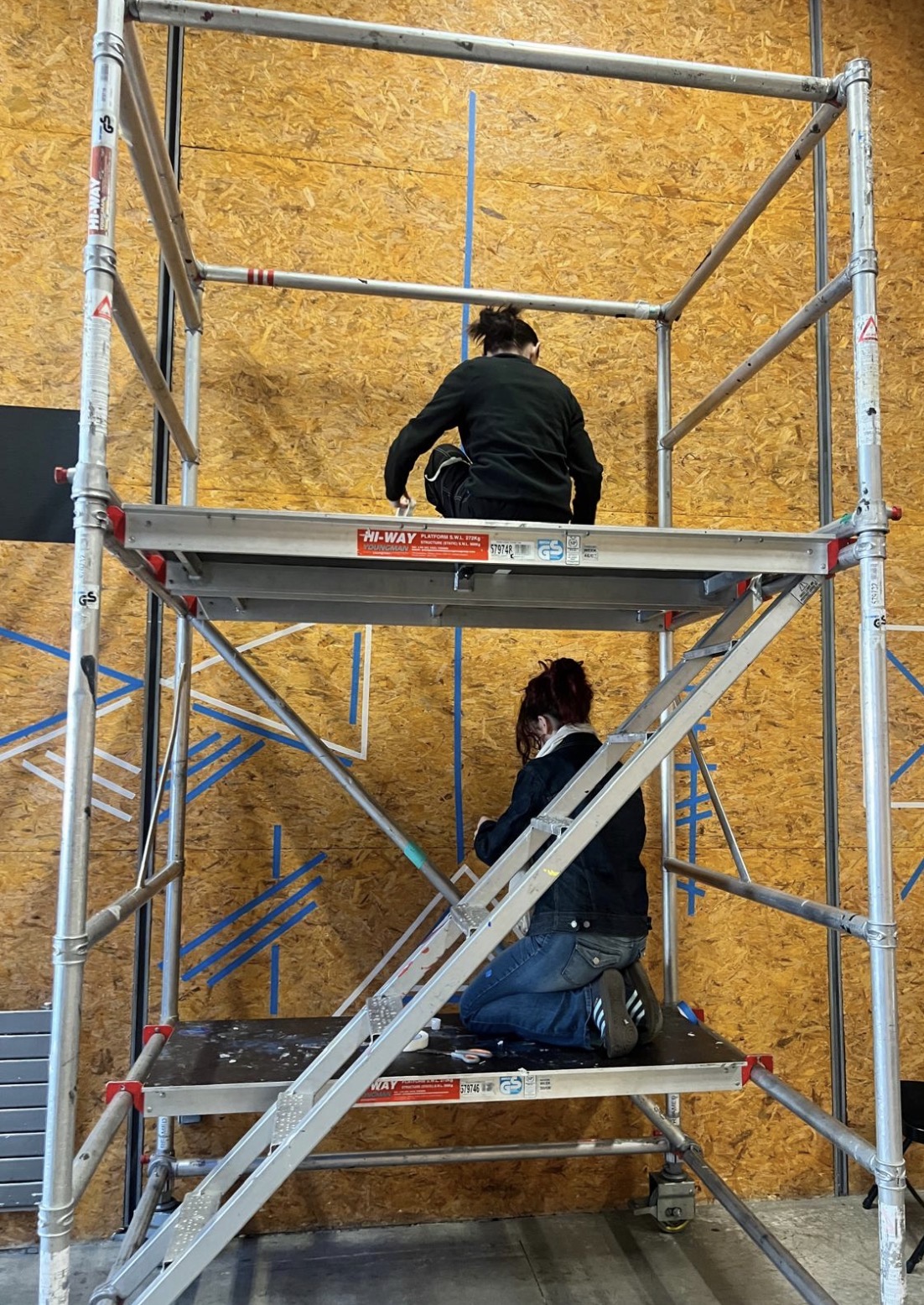 Freya and a collaborator painting a set wall from scaffolding
